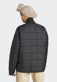 Femme portant un turban beige et une veste matelassée noire vue de dos, associée à un pantalon clair sur fond uni.