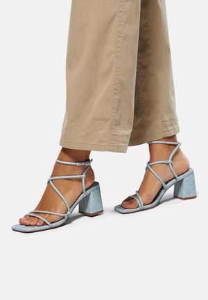 Sandale cu toc bloc cu bretele, din material textil de denin albastru deschis, cu vârful pătrat și multiple curele încrucișate. Tocul are o înălțime de aproximativ 5 cm.