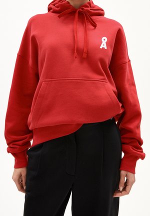 Personne portant un sweat à capuche rouge avec une poche avant et un logo blanc Å sur la poitrine, associé à un pantalon noir taille haute.