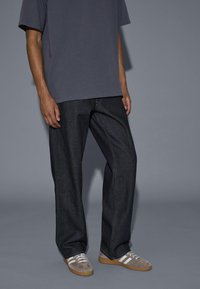 Person trägt dunkelblaue Jeans, graues T-Shirt und graue Wildledersneaker mit weißen Streifen, steht vor einem schlichten grauen Hintergrund.