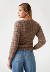 Pulover marron, pufos, cu o textură ribbed, mâneci lungi și decolteu rotund, purtat cu blugi din denim albastru. Detaliu subtil al logo-ului pe partea din spate.