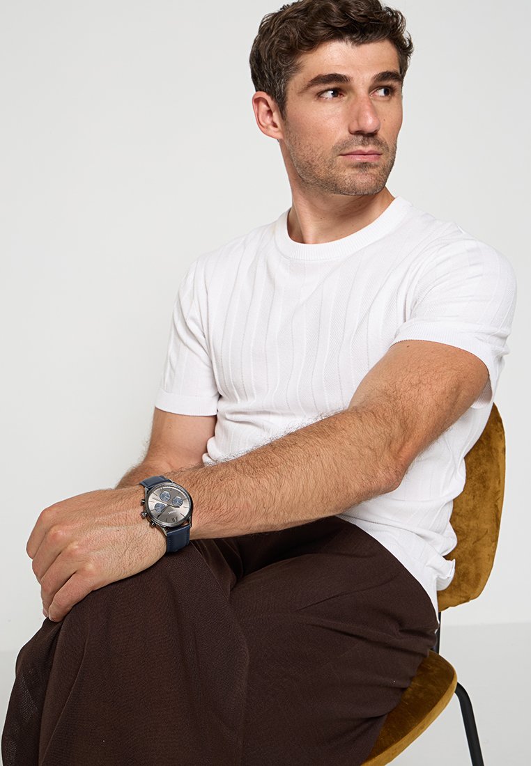 T-shirt blanc à rayures, pantalon ample marron, et une montre bleue à cadran rond. Modèle assis sur une chaise marron devant un fond clair.