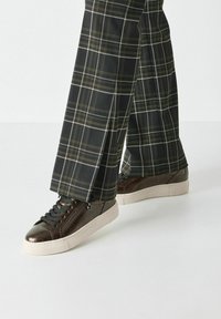Sneakers in pelle marrone con finitura testurizzata e lacci grigi, abbinati a pantaloni a quadri neri con linee bianche e gialle.