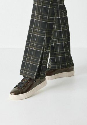 Sneakers in pelle marrone con finitura testurizzata e lacci grigi, abbinati a pantaloni a quadri neri con linee bianche e gialle.