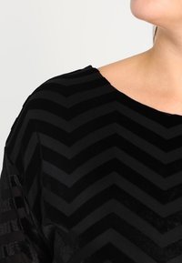 Black velvet top featuring a chevron pattern with alternating matte and shiny stripes. Wide neckline and short sleeves. Soft texture.