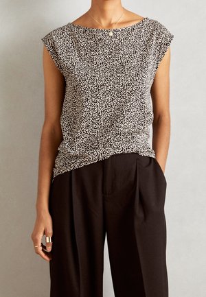 Vrouw draagt een zwart-wit patroon mouwloos topje, ingestopt in een hooggetailleerde zwarte geplooide broek, met ringen en een hangende ketting.