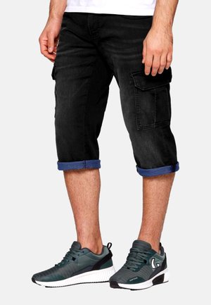 Fekete cargo shorts, feltekert szegéllyel, több zsebbel. Sötétzöld sportcipővel párosítva, texturált részletekkel és fehér talppal.