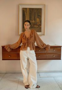 Vrouw in een bruin gerimpeld blouse en witte brede broek staat blootsvoets voor een houten lade en een ingelijste klassieke schilderij.