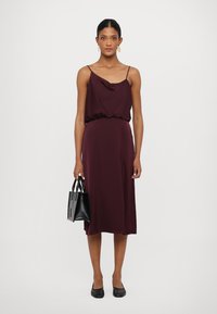Rochie midi în nuanța burgundă, cu un guler drapat și talie elastică, asortată cu o geantă mică neagră și balerini negri. Material neted, design minimalist.