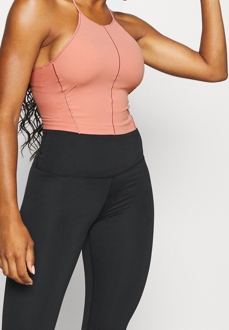 Femme portant un débardeur moulant rose poudré et un legging noir taille haute, levant son bras droit avec le coude fléchi.