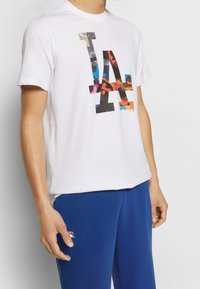T-shirt en coton blanc avec un logo "LA" camouflage multicolore. Associé à un pantalon de survêtement bleu, offrant une coupe décontractée et un design casual.