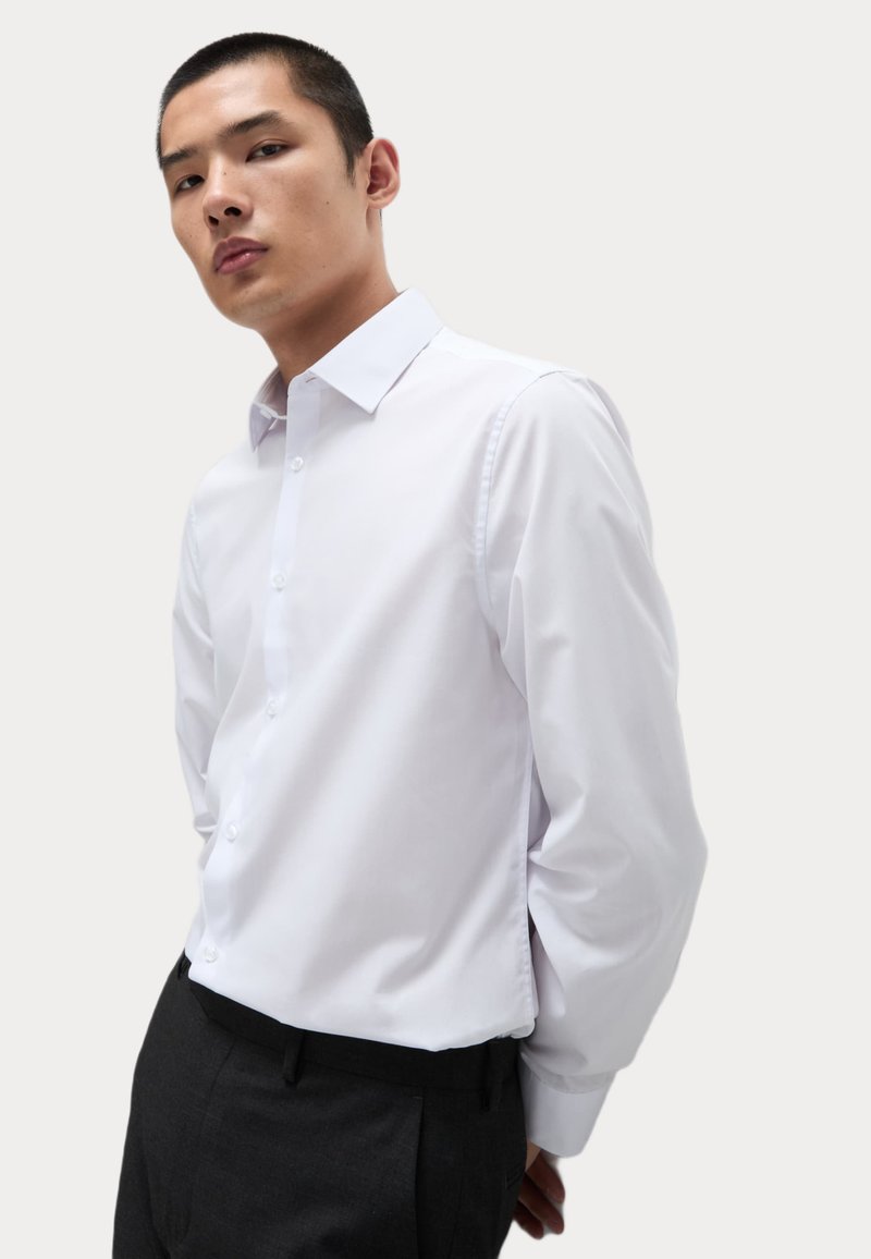 Camisa blanca de botones con cuello clásico y mangas largas, combinada con pantalones oscuros. Textura suave y ajuste entallado.