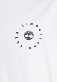 Timberland-logo med tre-symbol i senter, omgitt av teksten "timberland 1973" på hvit stoffbakgrunn.