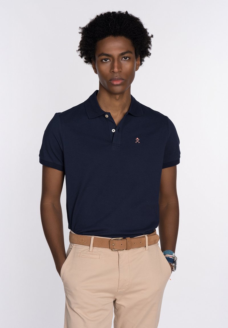 Polo de manga corta azul marino con cuello, que presenta un pequeño logo bordado rojo en el pecho y una textura suave de algodón. Cinturón beige combinado con pantalones color beige.
