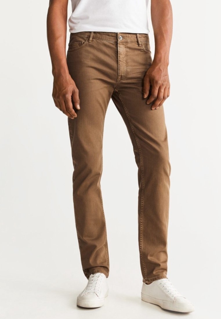 Homme portant un pantalon marron ajusté et des baskets blanches, debout devant un fond blanc uni.