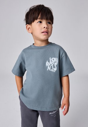 Junge mit kurzem dunklem Haar, der ein blau-graues AllSaints London T-Shirt und dunkle Hosen trägt, schaut leicht nach rechts.