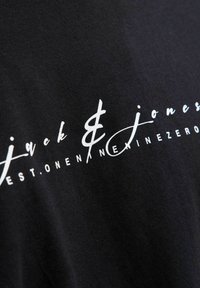 Juoda medžiaga su baltu stilizuotu tekstu "jack & jones" ir mažesniais didžiosiomis raidėmis žemiau, dalinai rašantis "EST. ONE NINE ZERO."