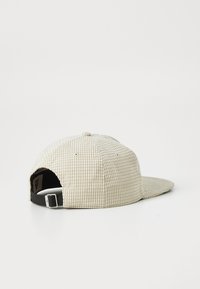 Boné de basebol em tecido texturizado bege, com fita ajustável preta e fivela prateada, mostrado de lado e atrás sobre um fundo branco.