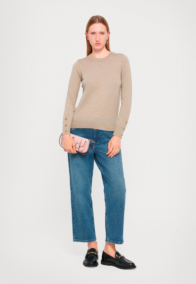 Beiger Strickpullover mit langen Ärmeln, goldenen Knopfdetails an den Manschetten, kombiniert mit blauen geraden Jeans und schwarzen Loafern. Hellrosa Clutch-Bag.