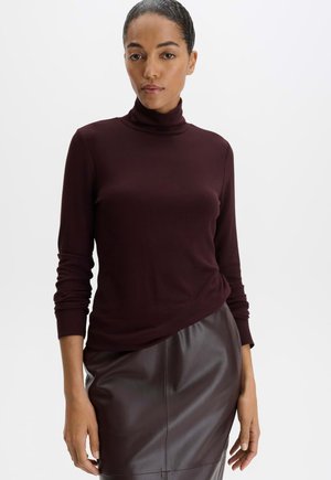 Femme portant un pull col roulé moulant bordeaux foncé et une jupe en cuir marron foncé sur un fond clair uni.