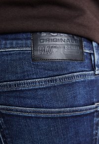 Jeans in denim blu scuro con cuciture visibili, dotati di un'etichetta in pelle nera testurizzata con la scritta "G-Star RAW" sulla cintura.