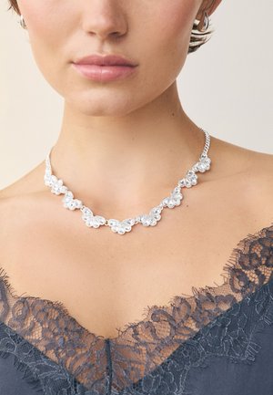 Silver necklace with floral design, featuring clear stones, a delicate chain, and decorative accents. Worn with a lace-trimmed garment.