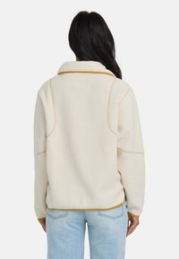 Frau mit langen, dunklen Haaren, die eine cremefarbene Fleecejacke mit senfgelbem Saum und hellblauen Jeans trägt und sich von der Kamera abwendet.