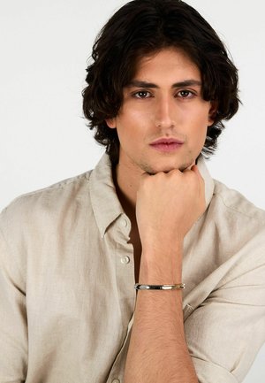 Bracciale d'argento al polso, indossato con una camicia di lino beige chiaro. Il bracciale presenta una superficie liscia e lucida e un design semplice.