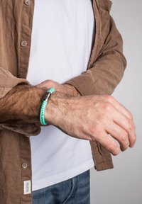 Bracciale intrecciato regolabile di colore turchese con chiusura metallica. Indossato su un polso, mostra un design testurizzato su una camicia di velluto a coste marrone.