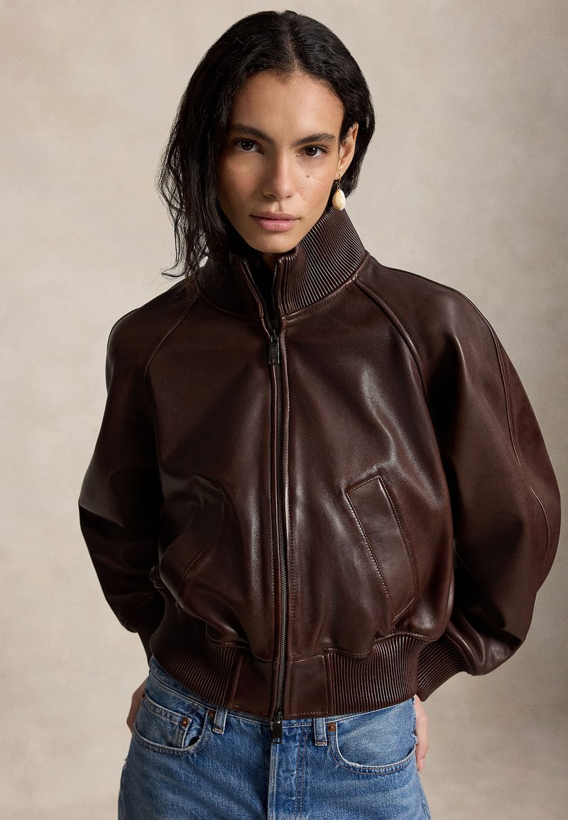 Γυναίκα με σκούρα μαλλιά φοράει σκούρο καφέ δερμάτινο μπουφάν με φερμουάρ, ριμπ γιακά και μανσέτες, μπλε τζιν και ένα μονό σκουλαρίκι που κρέμεται.
