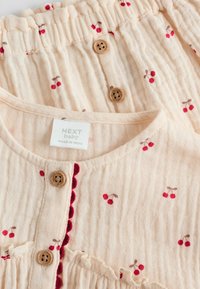 Lys beige bomuldsbabydress med kirsebærmønster, trækknapper og en bølget rød kant langs halsudskæringen og forsiden. Blødt, struktureret stof.
