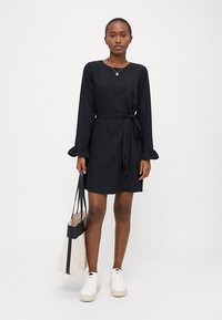 Μαύρο φόρεμα με μακριά μανίκια, με ρυάκια στους καρπούς, ζώνη στη μέση, μήκους μέχρι το γόνατο, συνδυασμένο με λευκά αθλητικά παπούτσια και μια δίχρωμη τσάντα ώμου.