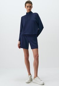 Completo sportivo blu navy composto da una felpa con mezza zip e orlo testurizzato, abbinata a pantaloni shorts. Completato con sneakers bianche. Tessuto liscio.