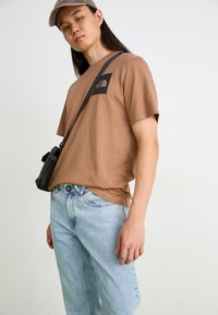 Bruine korte mouwen t-shirt met een zwart North Face-logo, gecombineerd met lichtblauwe jeans en een kleine zwarte schoudertas.