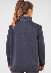 Pull noir en gris foncé avec un col haut, fabriqué en tissu doux. Présente un logo prominent sur le col et une coupe décontractée.