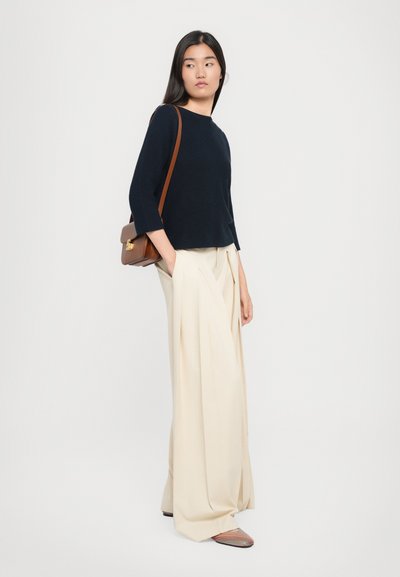 Navy strukturierter Pullover, weite cremefarbene Hose und gemusterte Slipper. Braune Umhängetasche mit einem goldenen Verschluss. Modell steht vor einem weißen Hintergrund.
