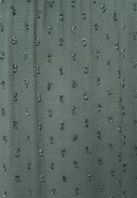 Tissu vert texturé présentant des motifs en relief et en pointillés sur toute sa surface. La surface globale est lisse avec un léger éclat.