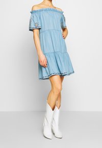 Vaaleansininen olkapäiltä avattava mekko, jossa on röyhelökaulus ja lyhyet hihat, sekä brodeerattu koristelu hihoissa ja kerroksellinen hame.