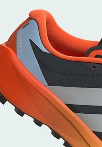 Gros plan sur une chaussure de sport orange, noire et grise avec une semelle texturée et le logo "TERREX" sur le col du talon.