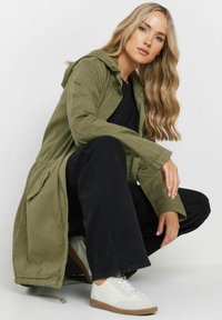 Lungo giubbotto verde oliva con cappuccio, in tessuto strutturato, abbinato a pantaloni neri e sneakers bianche. Dettagli notevoli includono un girovita con cordoncino e tasche laterali.