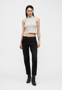 Femme tatouée portant un crop top à rayures sans manches avec col roulé, jean noir et bottines noires, debout devant un fond uni.