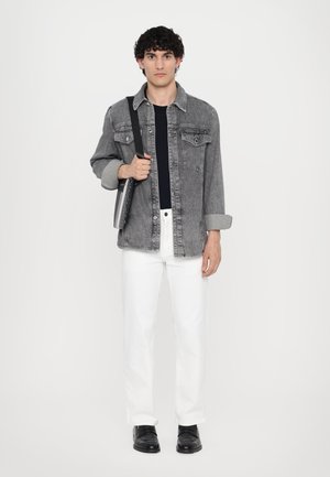 Chaqueta de mezclilla gris sobre una camiseta negra, pantalones blancos y zapatos negros. El modelo sostiene un bolso negro y está de pie contra un fondo liso.