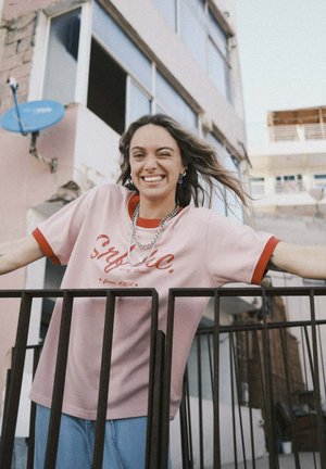 Różowa koszula z krótkim rękawem z czerwonymi akcentami i nadrukowanym napisem, zestawiona z jasnoniebieskimi dżinsami. Modelka opiera się o czarną poręcz.