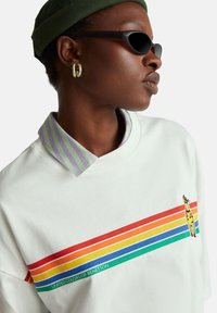 Biała bluza z nadrukiem w postaci wielokolorowych poziomych pasków, grafiką żyrafy oraz kołnierzem w jasnofioletowe i zielone paski.