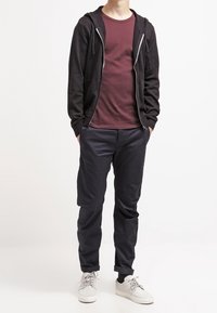 Svart zip-up hoodie över en vinröd t-shirt, kombinerad med mörkblå byxor och vita sneakers. Har en avslappnad passform och långa ärmar.