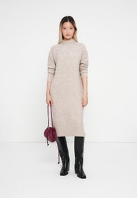 Abito a maglia in beige chiaro con maniche lunghe, collo alto e orlo a coste. Indossato con stivali neri con tacco e una borsa a tracolla bordeaux.