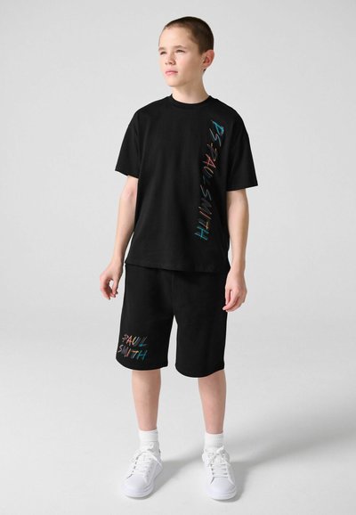 Garçon debout portant un t-shirt noir et un short avec le texte multicolore "Paul Smith", des baskets blanches et des chaussettes, sur un fond uni.