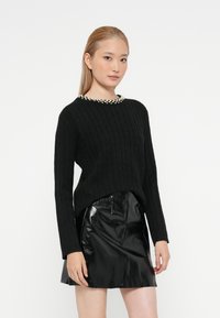 Jovem mulher com cabelo loiro liso veste um suéter preto de malha com decote de pérolas e uma saia preta de couro brilhante.