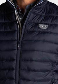 Marineblauwe gewatteerde vest met een quilted ontwerp, voorrits en logo patch. Kenmerken een hoge kraag en een gestructureerde voering. Duurzame stofafwerking.