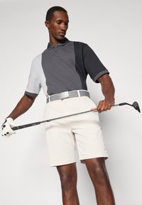 Polo cu blocuri de culoare gri și negru, cu mâneci scurte, asortat cu pantaloni scurți bej deschis. Jucătorul ține o crosieră și poartă un mănușă.